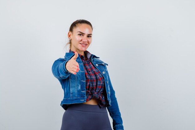 Mujer joven estirando la mano hacia adelante en camisa a cuadros, chaqueta de jean y aspecto lindo.