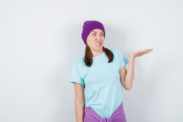 Mujer joven estirando la mano mostrando, mirándola haciendo muecas en camiseta azul, gorro morado y mirando acosado, vista frontal.