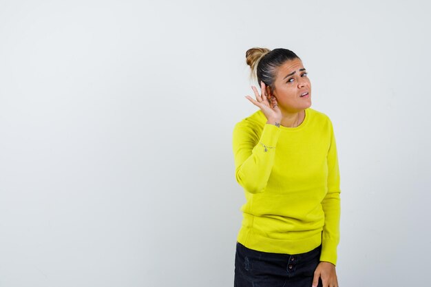 Mujer joven estirando las manos en cuestionamiento tomados de la mano cerca de la oreja para escuchar algo en suéter amarillo y pantalón negro y mirando enfocado