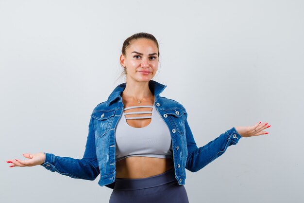 Mujer joven estirando las manos a un lado en la parte superior de la cosecha, chaqueta, pantalones y mirando confiado, vista frontal.