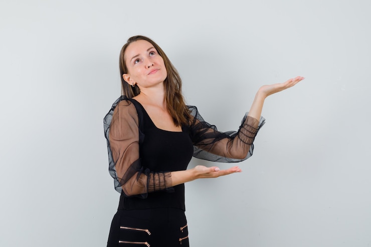 Foto gratuito mujer joven estirando las manos presentando algo en blusa negra y pantalón negro y mirando feliz, vista frontal.