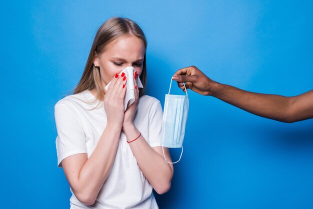 Mujer joven estornuda mientras la mano da máscara médica aislada en la pared azul