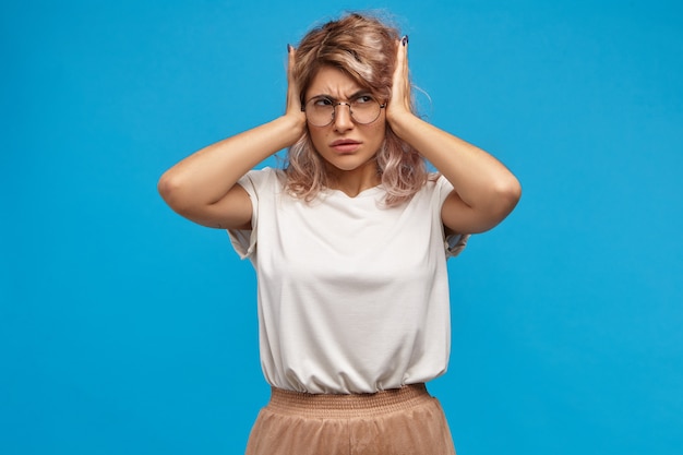 Mujer joven estresada irritada en gafas redondas que cubren las orejas, siendo molestada con mucis fuerte