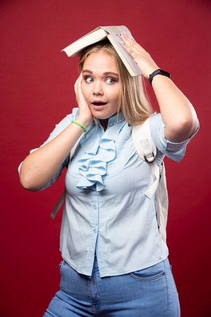 Mujer joven estudiante rubia sostiene su libro en la cabeza y parece cansada y confundida.