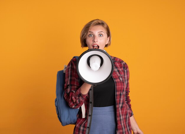Mujer joven estudiante sorprendida con auriculares con mochila habla en el altavoz aislado en la pared naranja