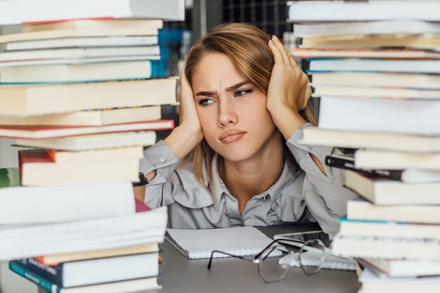 Mujer joven estudiante universitaria infeliz en una biblioteca, posando con gafas y libros.