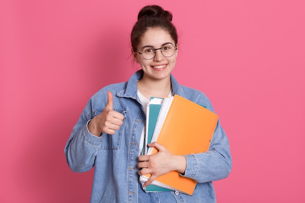 Mujer joven estudiante vistiendo chaqueta vaquera y anteojos, sosteniendo carpetas de colores y mostrando el pulgar hacia arriba en rosa