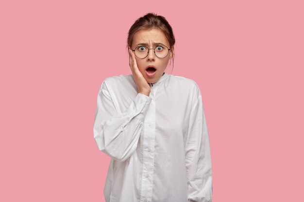 Mujer joven estupefacta con gafas posando contra la pared rosa