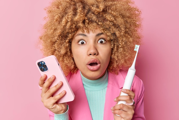 Mujer joven estupefacta y sorprendida mantiene la boca abierta por el asombro sostiene el teléfono móvil y el cepillo de dientes eléctrico limpia los dientes regularmente descubre algo horrible aislado sobre la pared rosa del estudio