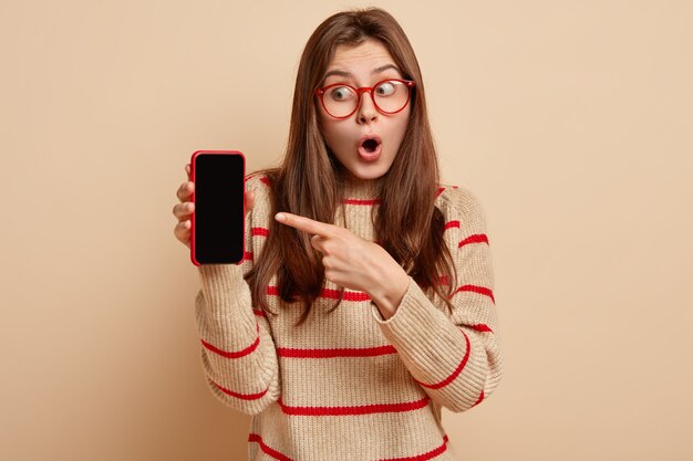 Mujer joven europea sorprendida abre la boca de asombro, apunta al teléfono móvil con pantalla en blanco para la plantilla de contenido, diseño, usa un jersey beige con rayas rojas, aislado sobre una pared marrón