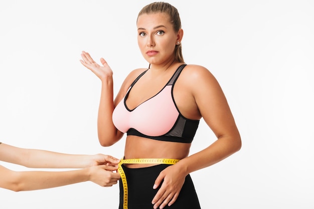 Mujer joven con exceso de peso en la parte superior deportiva sorprendida mirando a la cámara mientras mide la cintura sobre fondo blanco.