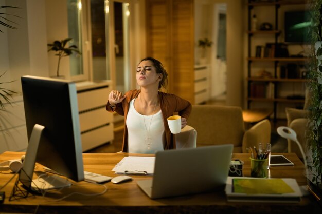 Mujer joven exhausta estirándose mientras trabaja tarde en la noche en casa