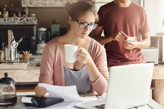 Mujer joven con expresión concentrada mirando la pantalla de la computadora portátil abierta, sosteniendo papel y una taza de café en sus manos mientras calcula los gastos domésticos