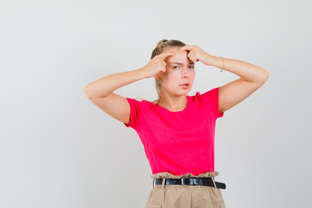 Mujer joven exprimiendo su grano en la frente en camiseta, pantalón, vista frontal.