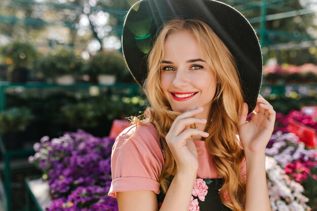 Mujer joven extasiada con ojos claros posando cerca de flores. Linda mujer europea con sombrero escalofriante en invernadero.