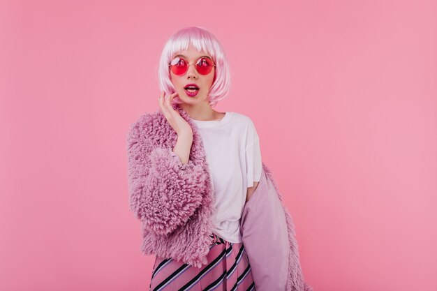 Mujer joven extática en chaqueta mullida divertida posando en la pared rosa. Retrato de interior de increíble jocund niña caucásica en periwig