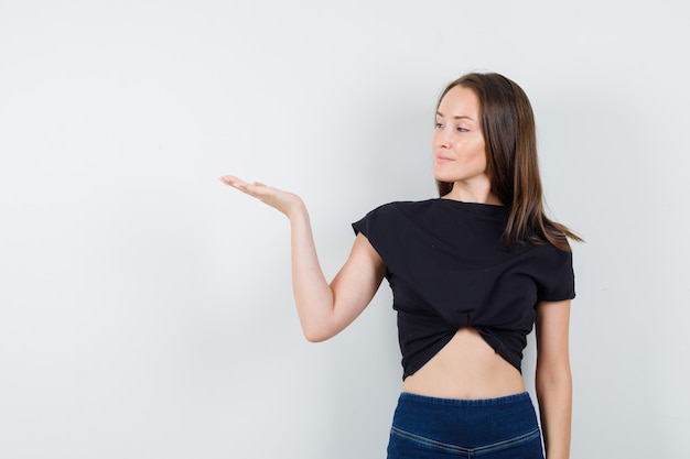 Mujer joven extendiendo la palma a un lado en blusa negra, pantalones y mirando confiado