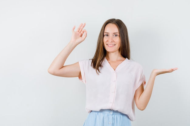Mujer joven extendiendo la palma a un lado con gesto ok en camiseta, falda y mirando positivo, vista frontal.