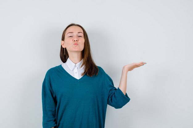 Mujer joven extendiendo la palma a un lado, haciendo pucheros con los labios en suéter sobre la camisa blanca y mirando pacífica, vista frontal.