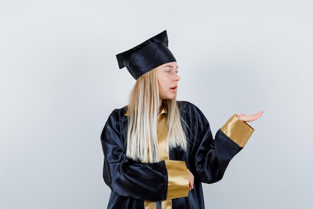 Foto gratuita mujer joven extendiendo la palma a un lado en uniforme de posgrado y mirando enfocado.