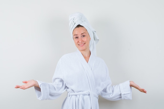 Mujer joven extendiendo las palmas a un lado en bata de baño blanca, toalla y mirando alegre. vista frontal.