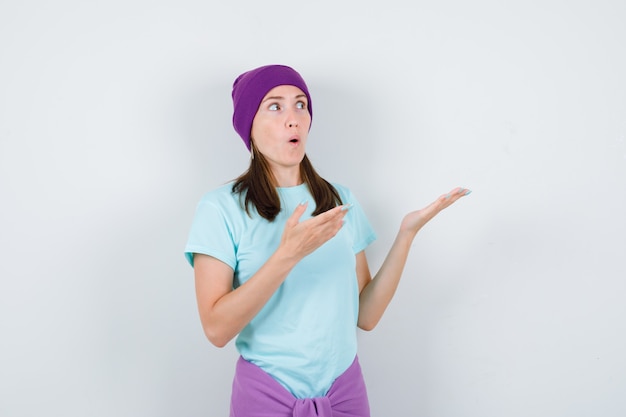 Mujer joven extendiendo las palmas a un lado, manteniendo la boca abierta en camiseta azul, gorro morado y mirando sorprendido. vista frontal.