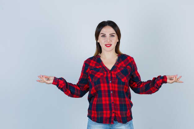 Mujer joven extendiendo las palmas hacia los lados en camisa a cuadros y mirando alegre. vista frontal.