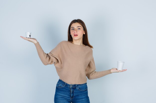 Mujer joven extendiendo las palmas con vasos de papel en suéter, jeans y mirando sorprendido, vista frontal.