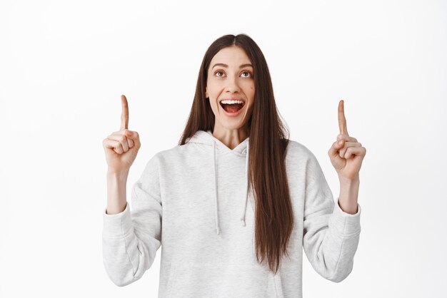 Mujer joven fascinada revisando el texto promocional señalando y mirando asombrada por el logotipo de la pancarta oferta de trato súper genial de pie en una sudadera con capucha contra fondo blanco