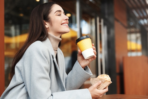 Mujer joven feliz en abrigo gris comiendo una rosquilla en una cafetería.