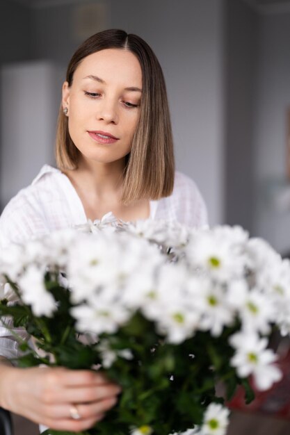 Mujer joven feliz y alegre de blanco arreglando flores blancas en casa en la cocina