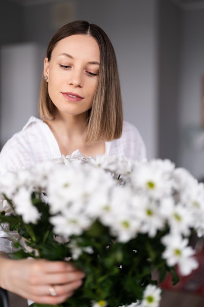 Mujer joven feliz y alegre de blanco arreglando flores blancas en casa en la cocina