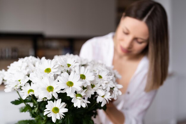 Mujer joven feliz y alegre de blanco arreglando flores blancas en casa en la cocina