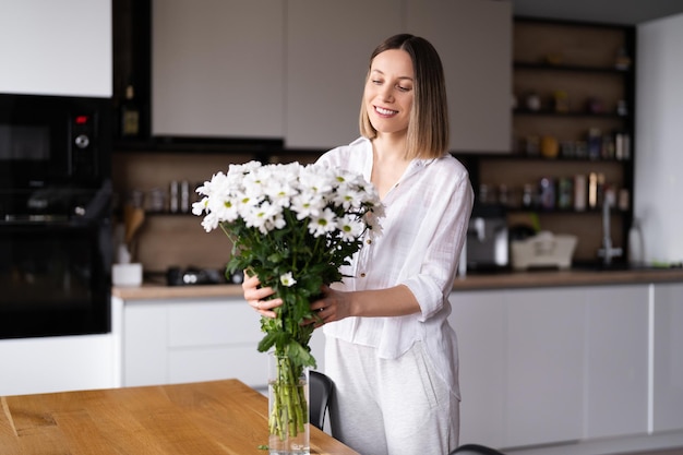 Mujer joven feliz y alegre de blanco arreglando flores blancas en casa en la cocina