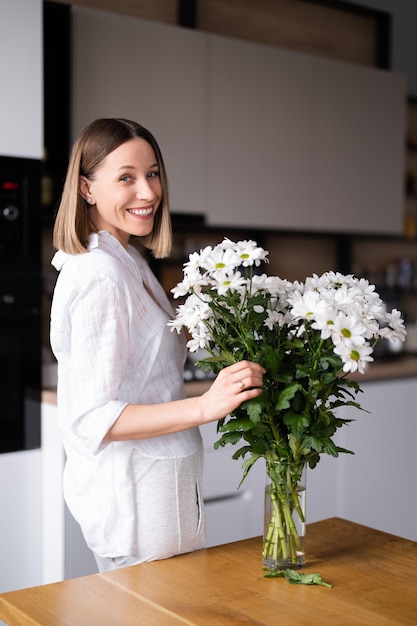 Mujer joven feliz y alegre de blanco arreglando flores blancas en casa en la cocina