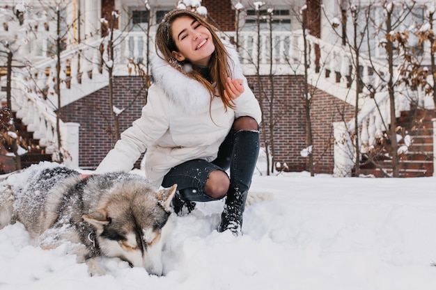 Mujer joven feliz alegre divirtiéndose con lindo perro husky en la nieve en la calle. Estado de ánimo alegre, invierno nevando, adorables mascotas en casa, verdadera amistad.