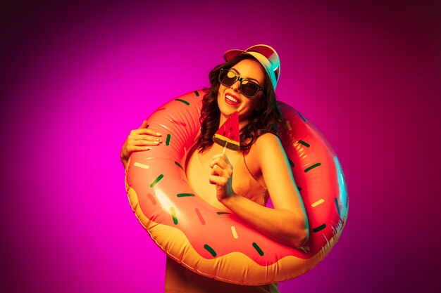 Mujer joven feliz en un anillo de playa de goma, gorra roja y gafas de sol con un caramelo en neón rosa de moda