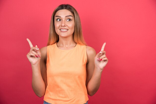 Mujer joven feliz apuntando su dedo hacia arriba en la pared roja.