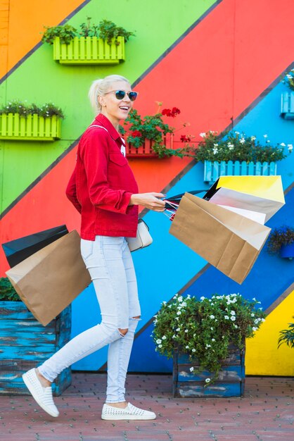 La mujer joven feliz con los bolsos acerca a la pared