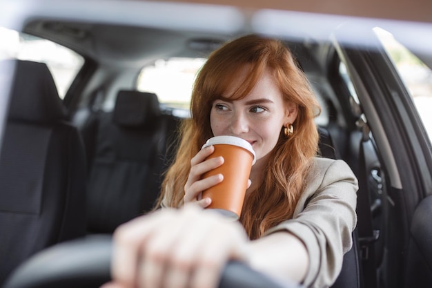 Mujer joven feliz con café para ir a conducir su automóvil Mujer tomando un café mientras conduce un automóvil Mujer joven tomando café mientras conduce su automóvil Pelirroja atractiva conduce un automóvil