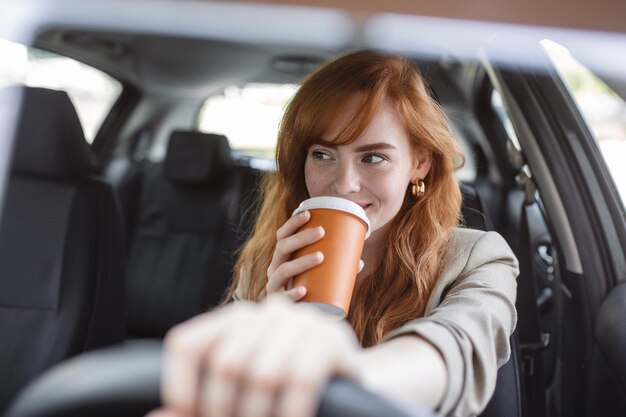 Mujer joven feliz con café para ir a conducir su automóvil Mujer tomando un café mientras conduce un automóvil Mujer joven tomando café mientras conduce su automóvil Pelirroja atractiva conduce un automóvil