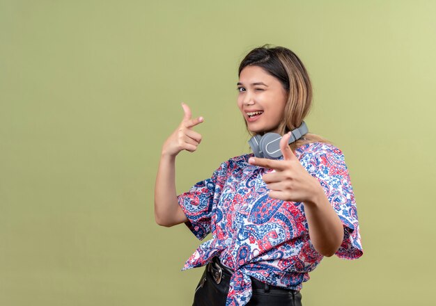 Una mujer joven feliz en camisa estampada de paisley usando audífonos apuntando con el dedo índice mientras mira en una pared verde