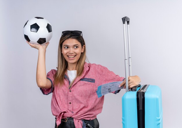 Una mujer joven feliz con camisa roja y gafas de sol mirando de lado mientras sostiene la bola con boletos de avión y maleta azul sobre una pared blanca