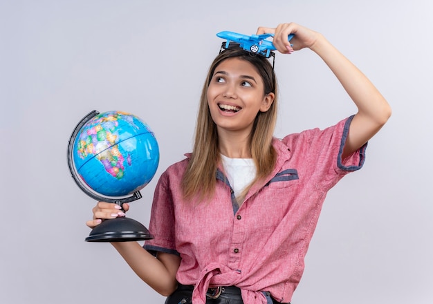 Foto gratuita una mujer joven feliz con camisa roja con gafas de sol sosteniendo un globo mientras volaba un avión de juguete sobre una pared blanca