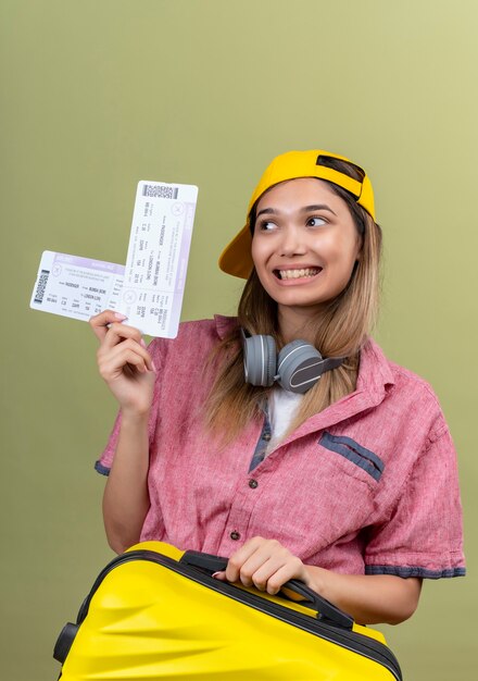 Una mujer joven feliz con camisa roja y gorra de béisbol amarilla que muestra los boletos de avión mientras lleva una maleta amarilla en una pared verde
