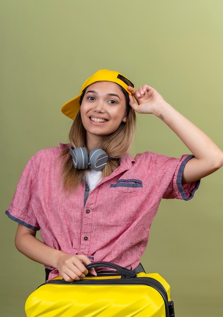 Una mujer joven feliz con camisa roja y gorra de béisbol amarilla sosteniendo una maleta amarilla mientras toca su sombrero en una pared verde