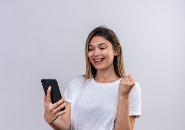 Una mujer joven feliz en camiseta blanca mirando el teléfono móvil con los puños cerrados sobre una pared blanca