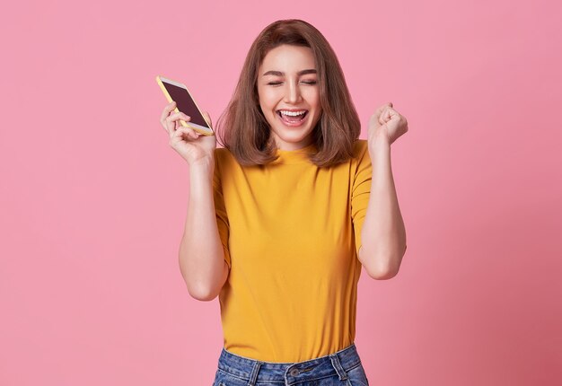 Mujer joven feliz celebrando con teléfono móvil aislado sobre fondo rosa.