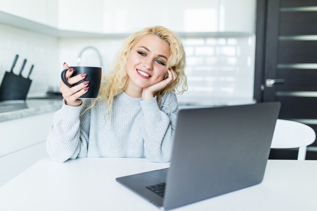 Mujer joven feliz en la cocina leyendo las noticias en su computadora portátil