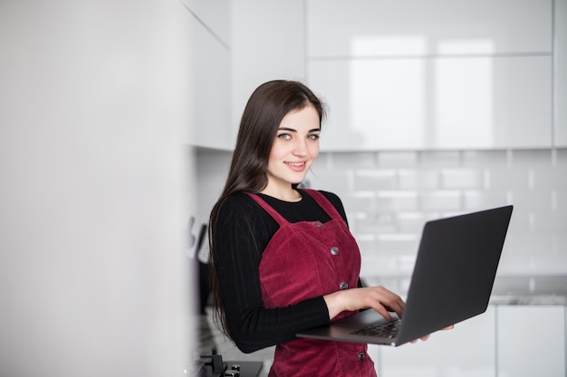 Mujer joven feliz en la cocina leyendo las noticias en su computadora portátil